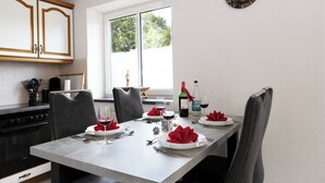 Mesa para cuatro servida con servilletas rojas, copas de vino y platos. Se ve una ventana con vistas a árboles y un reloj en la pared.