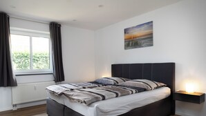 Un dormitorio con cama, ventana con cortinas, un cuadro colgado en la pared y una mesita de noche con lámpara.