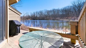 A patio with a round table and chairs, overlooking a calm lake with trees in the background. - Abbey Springs Condo with Golf Cart Pools Private Beach and Waterfront Deck (Fontana)