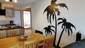 A dining area with a wooden table and chairs, a fruit bowl, and a wall mural of palm trees. - Gite de la Digue **** Wissant Cote d'Opale (Wissant)