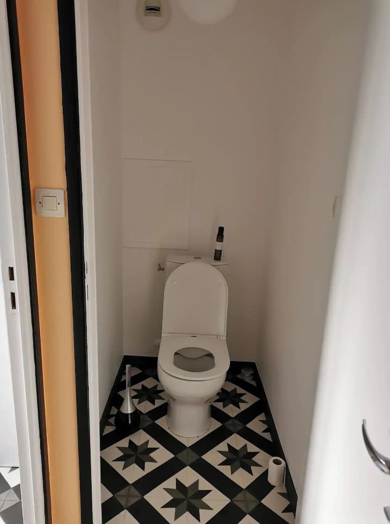 A small, narrow bathroom with a toilet, a wall-mounted flush handle, and a patterned floor.