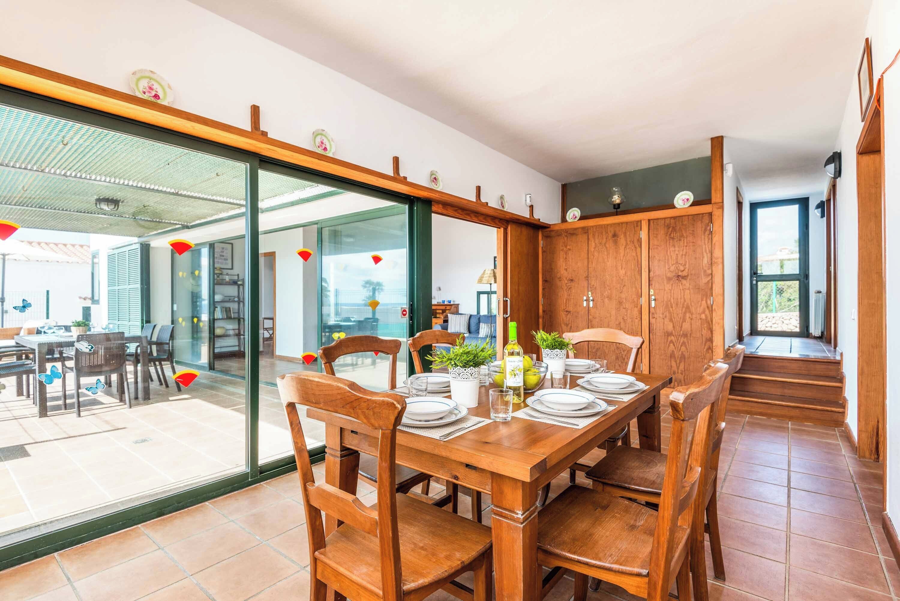 A dining area with a wooden table set for four, surrounded by wooden chairs. There are potted plants on the table, and a view through the glass door into another room with a dining table and chairs.