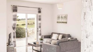 Ein modernes Wohnzimmer mit einem grauen Sofa, einem Couchtisch und einem Fernseher auf einem Schrank. Ein Glastür führt in einen Garten.