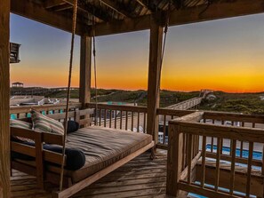 A wooden deck with a swing bed overlooking a beach at sunset. - Blue Wave - KDH MP 7.5, Oceanfront, Heated Pool, Hot Tub, Pool Table, Pets OK, Flex Stays (Kill Devil Hills)