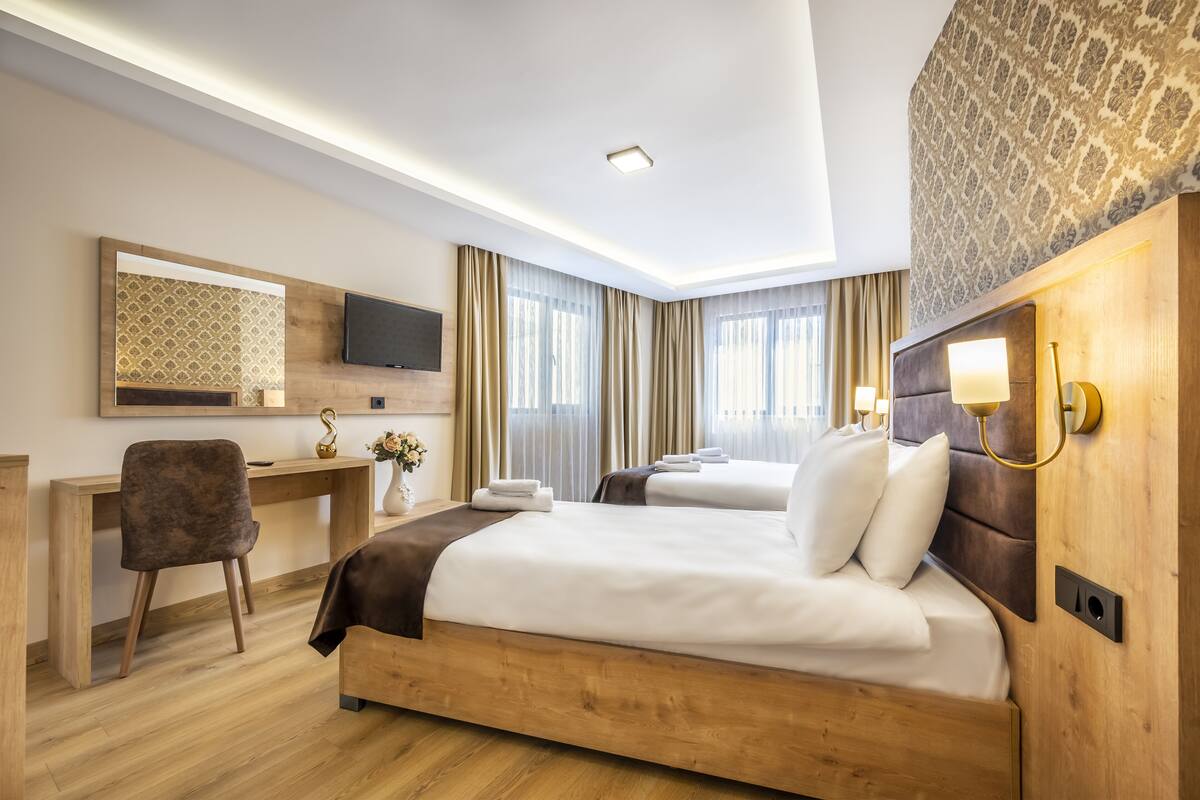A hotel room with a large bed, a desk, a chair, a TV, and a wall-mounted light fixture.