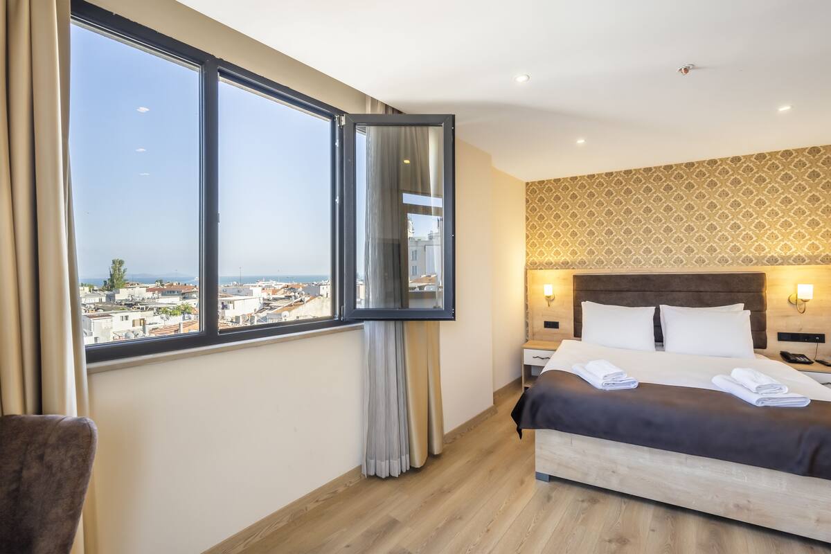 A hotel room with a large window, a bed with neatly folded towels, a wooden floor, and a patterned wall.