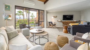 A spacious living room with a large sofa, a coffee table, and a TV. There is a balcony with outdoor furniture and palm trees.