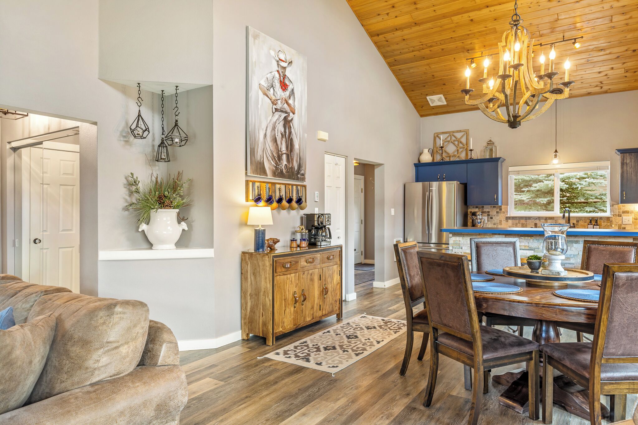 A spacious living room with a large couch, a wooden dining table set for four, and a kitchen area with blue cabinets and a chandelier.