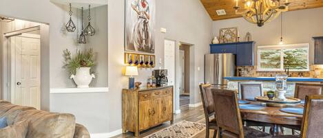 A spacious living room with a large couch, a wooden dining table set for four, and a kitchen area with blue cabinets and a chandelier.