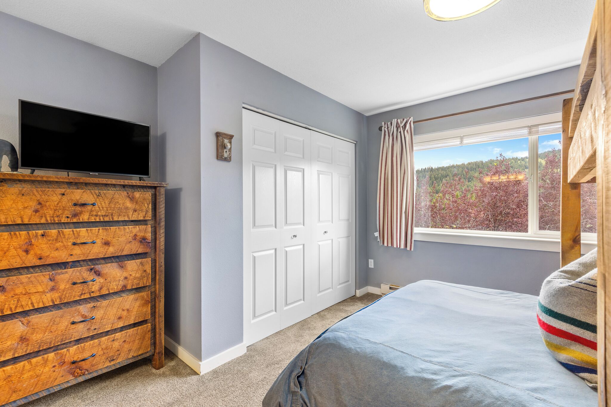 A bedroom with a bed, a dresser, a television, and a window with a view of trees.