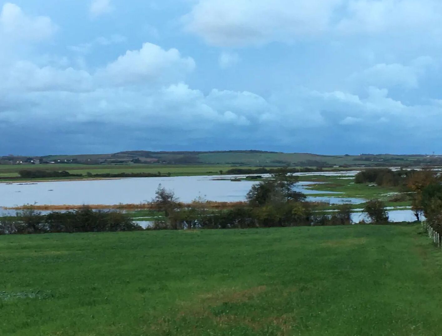 Un paysage rural avec un vaste champ ouvert, une étendue d’eau et des collines lointaines sous un ciel nuageux.