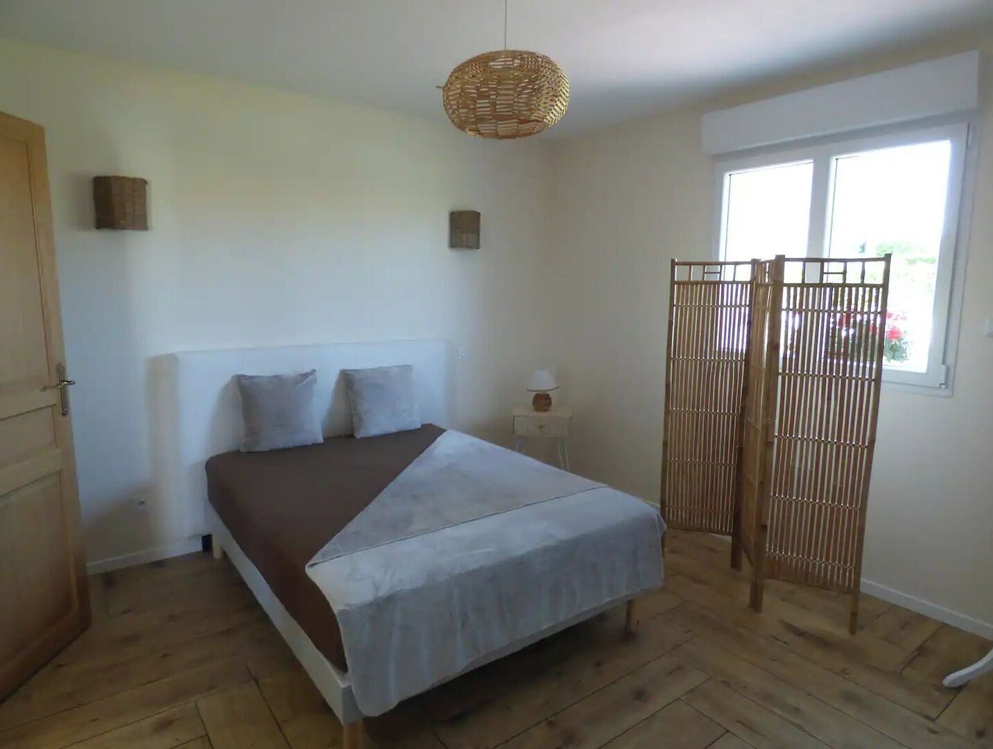 A bedroom with a bed, a bedside table, a lamp, a window, and a wooden floor.