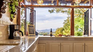A kitchen with a view of mountains and greenery, a coffee machine, and a wine bottle on the counter. - Ocean View 🌅Infinity Pool Villa & Hot Tub !CLOSE TO BEACH! (Jaco Beach)