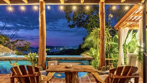 A wooden deck with two chairs and a table, overlooking a pool and a scenic view of hills at dusk. - Ocean View 🌅Infinity Pool Villa & Hot Tub !CLOSE TO BEACH! (Jaco Beach)