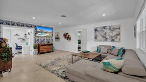 A spacious living room with a beige sofa, a coffee table, and a TV mounted on the wall. There is a wooden cabinet and a plant in the corner.