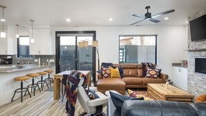 A modern living room with a sofa, a black leather couch, a dining area with a table and bar stools, a fireplace, and a large window. - Downtown Retreat | Steps to Bike Path &Restaurants (Moab)