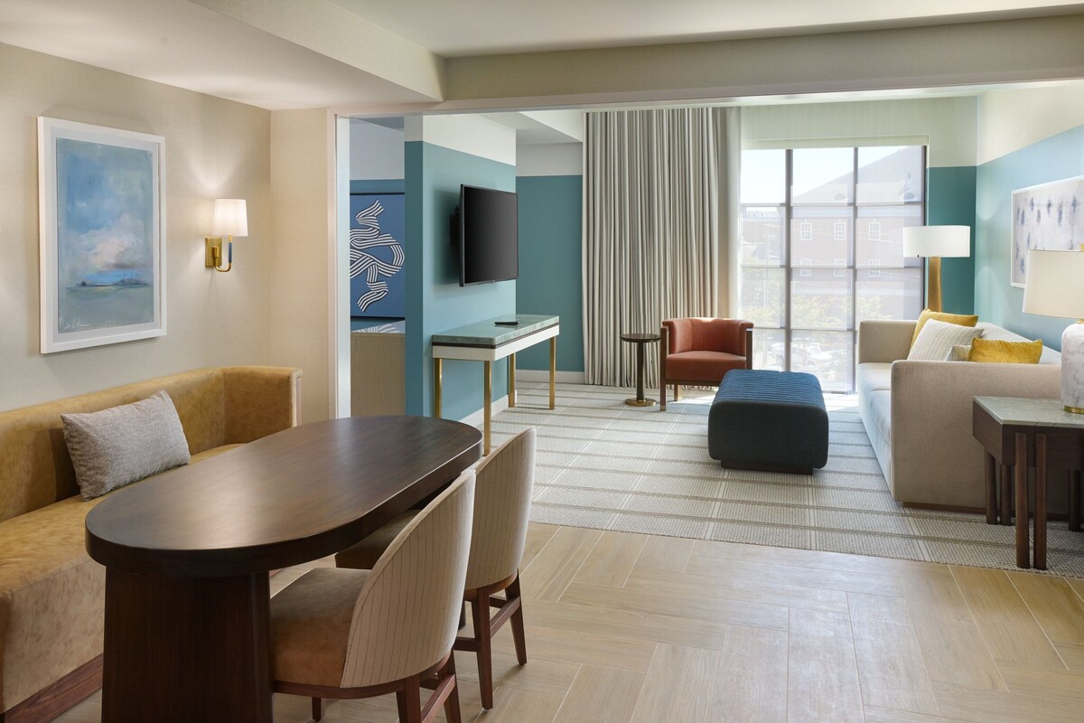 A hotel room with a dining area, a sofa, a TV, and a large window with curtains.