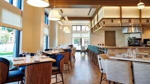 A modern restaurant with wooden tables and chairs, large windows, and hanging pendant lights.