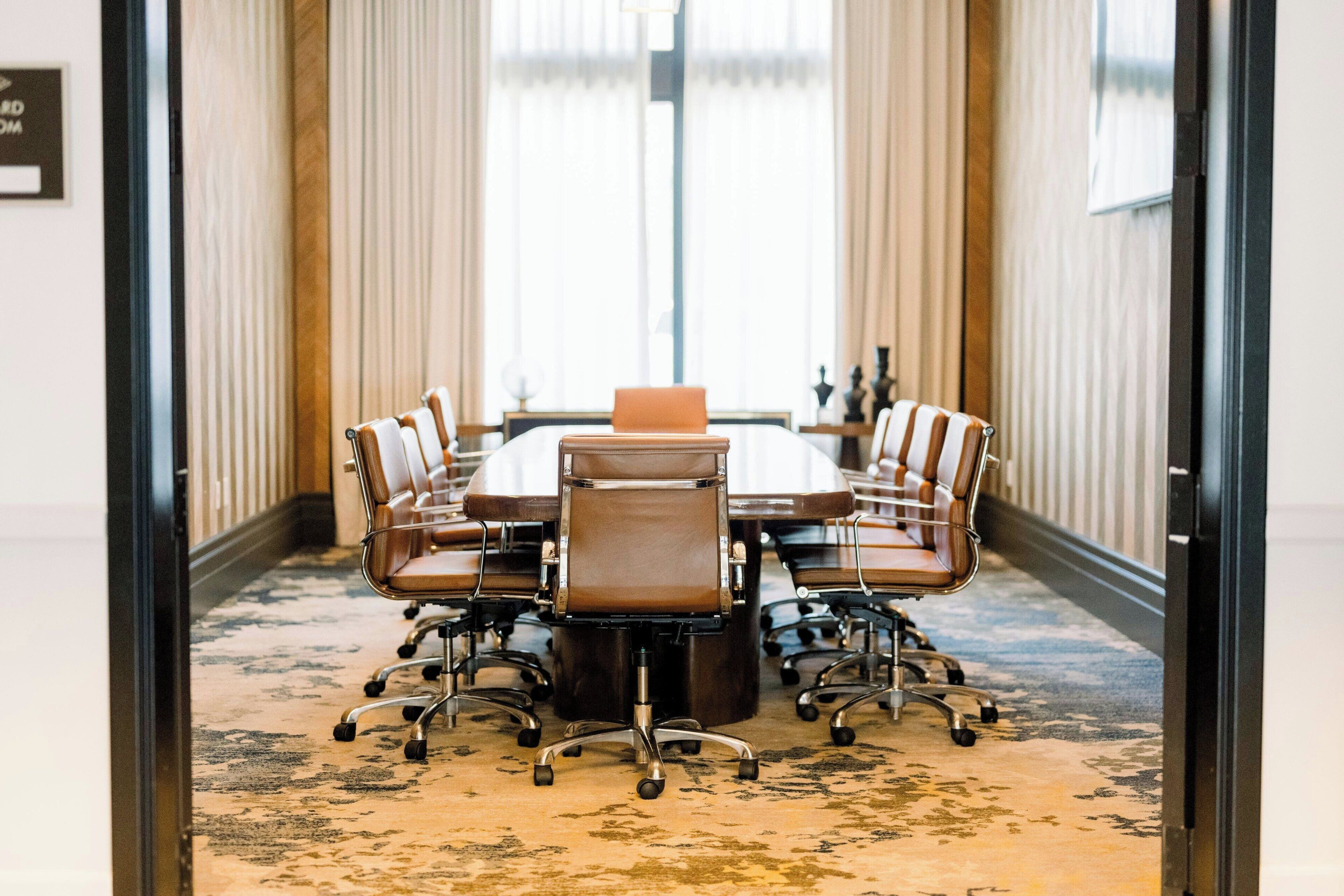 A conference room with a large table and six chairs, a carpeted floor, and a view through a glass door.