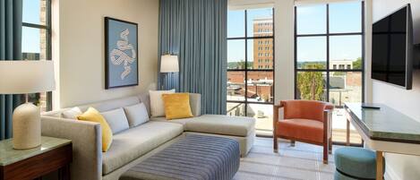 Una habitación de hotel moderna con sofá modular, una mesita, una silla, televisor y ventanas amplias con vistas a la ciudad.