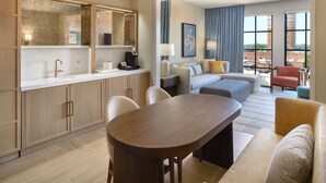 A modern hotel room with a kitchen area, a dining table, a sofa, and a view of the city.