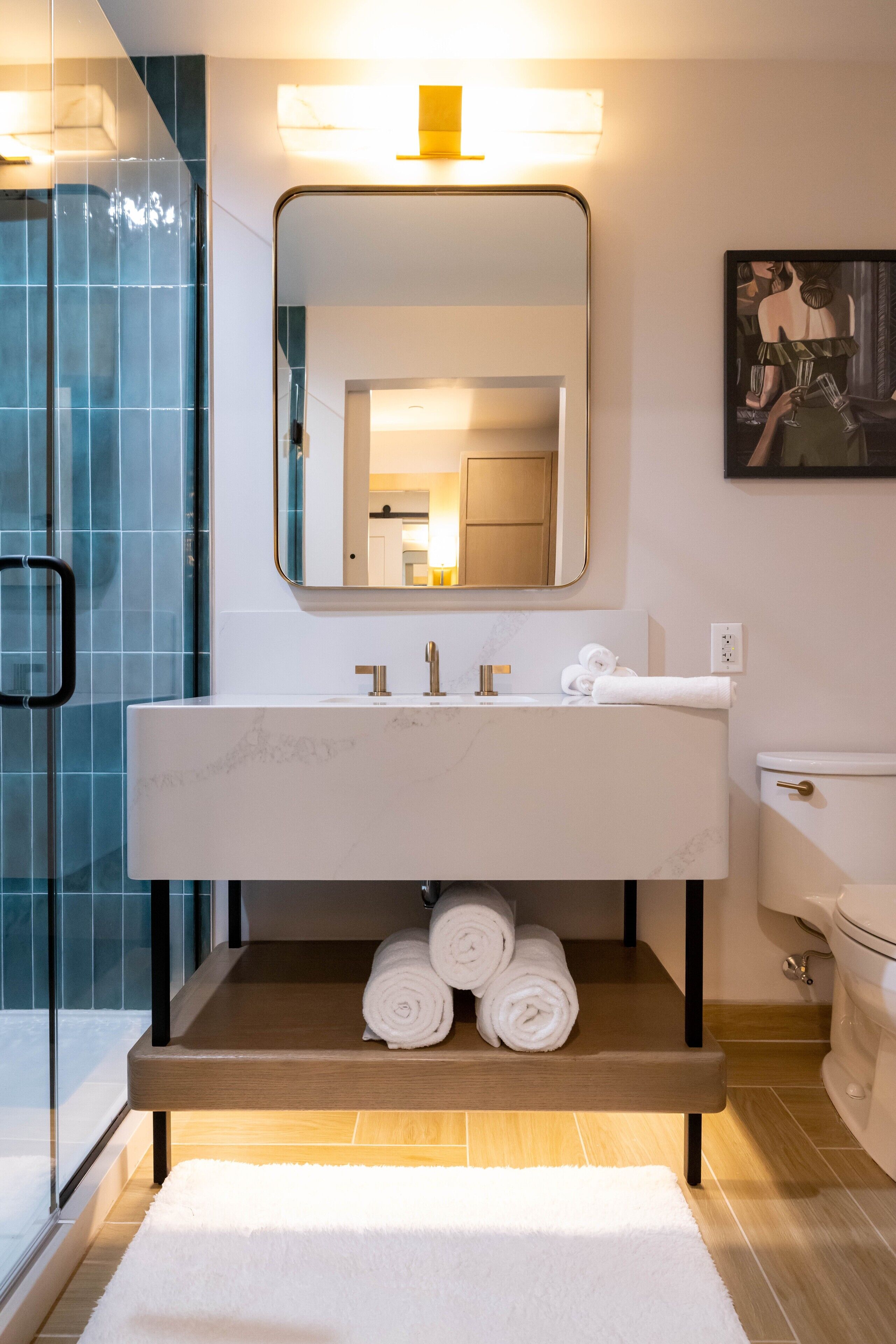 Een moderne badkamer met een witte wastafel, een spiegel en een toilet. Handdoeken liggen op een plank en er is een douchecabine met glazen wanden.