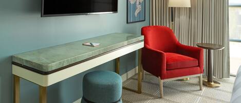 A hotel room with a flat-screen TV mounted on the wall, a red armchair, a small round table, a blue ottoman, and a patterned carpet.
