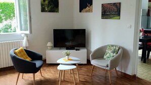 Ein Wohnzimmer mit Fernseher, zwei Sesseln, einem kleinen Tisch mit Vase und Kunstwerken an den Wänden.
