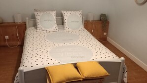 Ein ordentlich bezogenes Bett mit Punkteoptik und zwei Kissen, ein kleiner Nachttisch mit Lampe sowie eine Holzbank am Fußende.