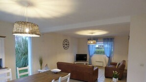 A living room with a wooden dining table, white chairs, a brown sofa, and a TV.