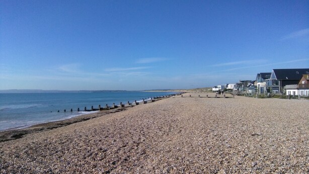 A pebbled beach with a wooden jetty, houses in the background, and a clear blue sky.