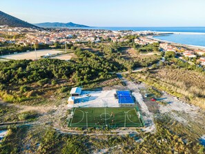 Vista aérea de uma vila costeira, com quadra esportiva cercada por vegetação e edifícios residenciais.