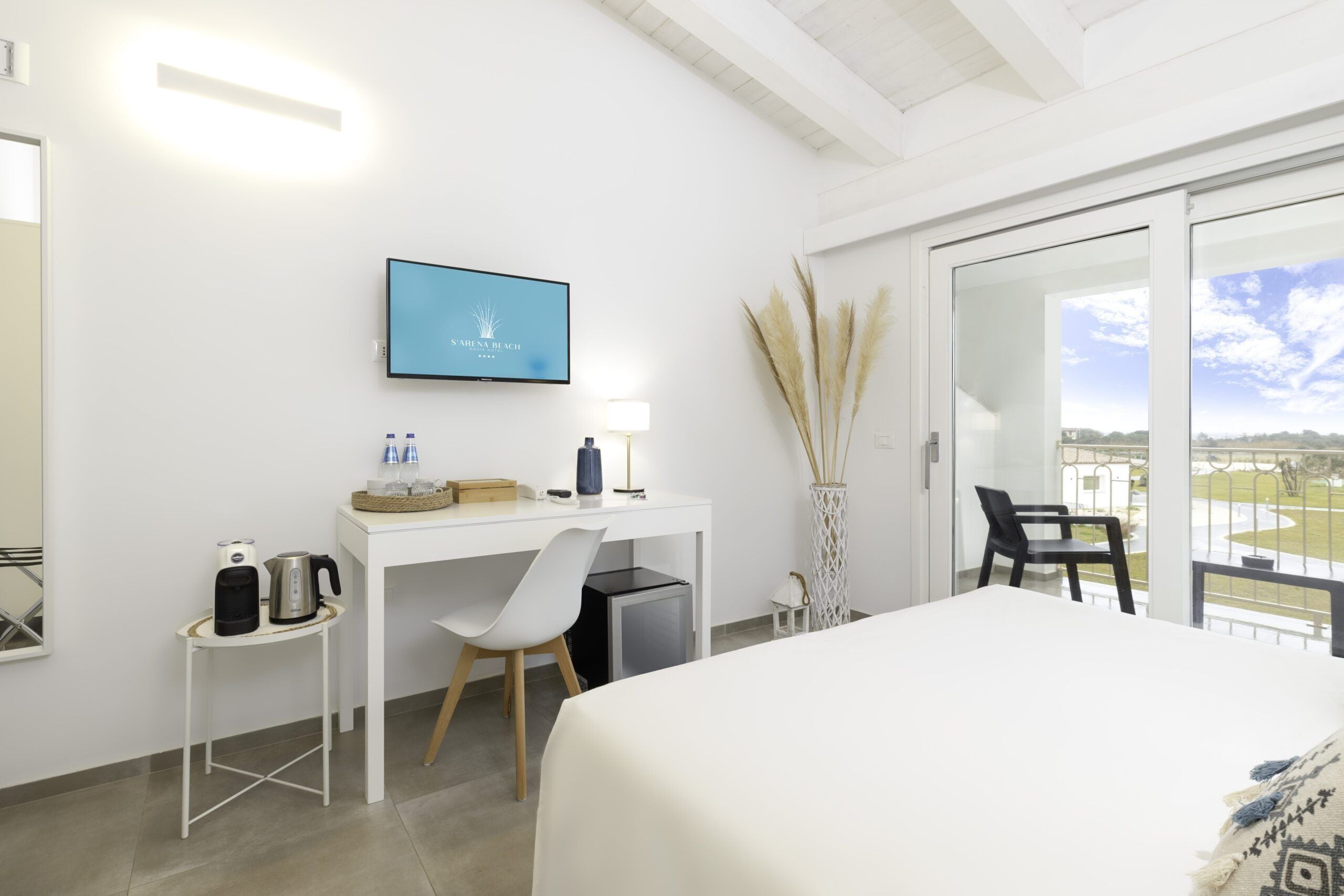 A hotel room with a bed, a desk with a computer, a chair, a small table with a kettle, a lamp, and a view of the outside.