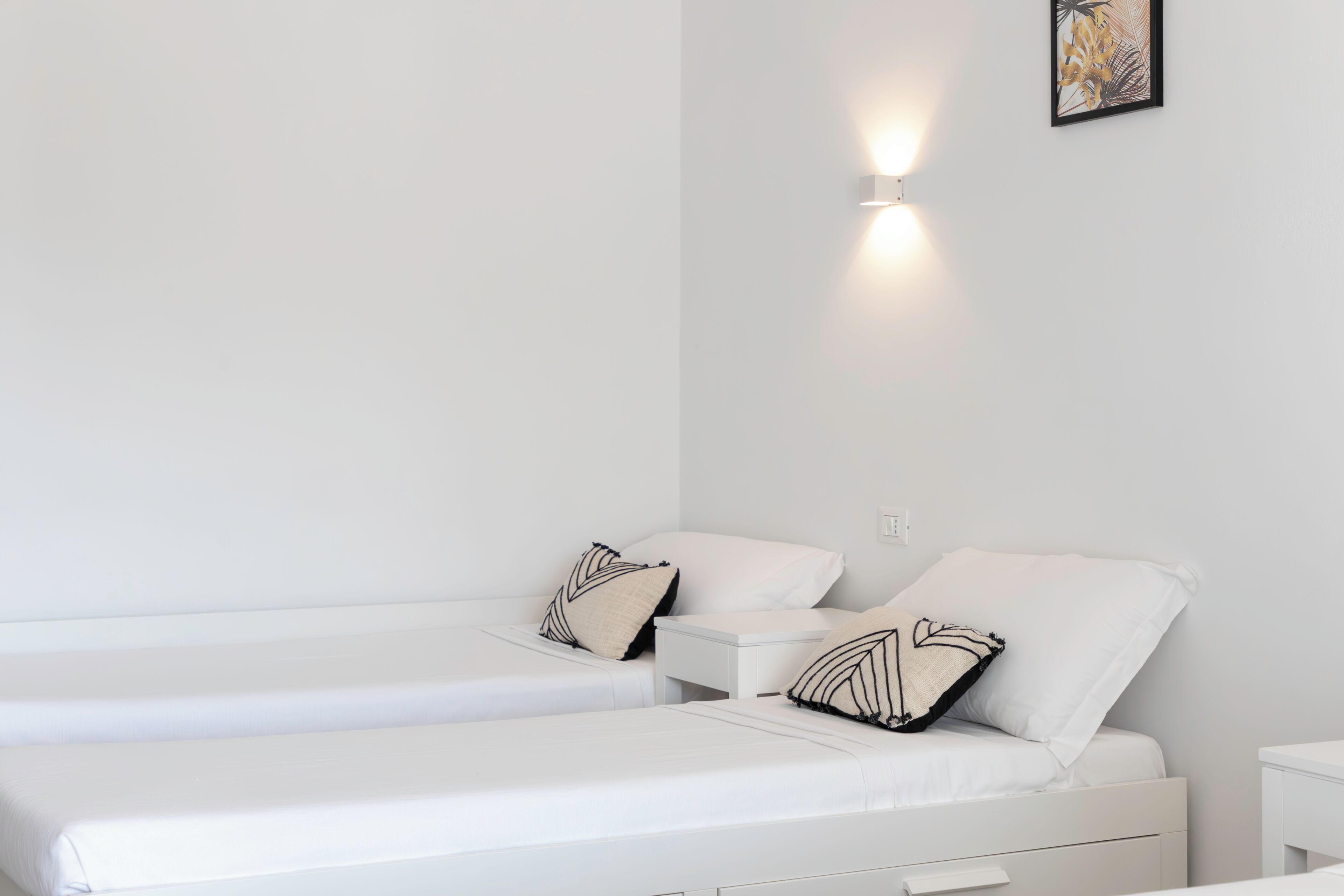 Un dormitorio minimalista con dos camas, ropa de cama blanca y una lámpara fijada a la pared.