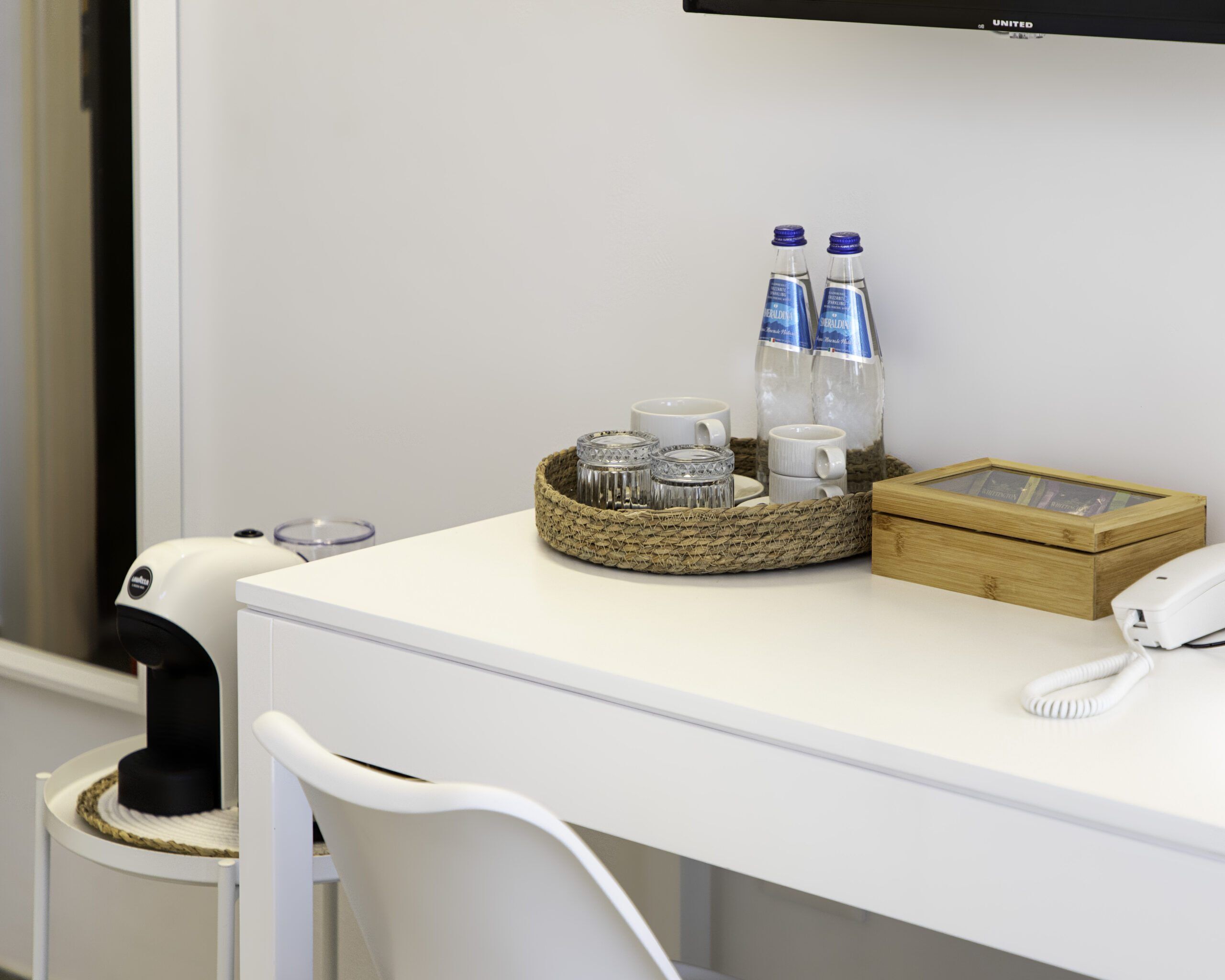 Une chambre d’hôtel avec un bureau blanc, un téléphone, un panier contenant des bouteilles d’eau et une boîte en bois.