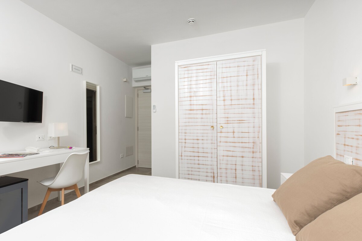A modern hotel room with a white bed, a desk with a chair, a television, and a wardrobe with a grid pattern.