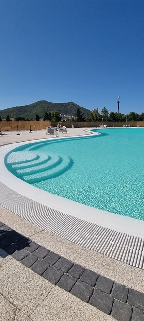 Piscina com característica de água azul cristalina, rodeada por um deck ladrilhado e algumas cadeiras de descanso.