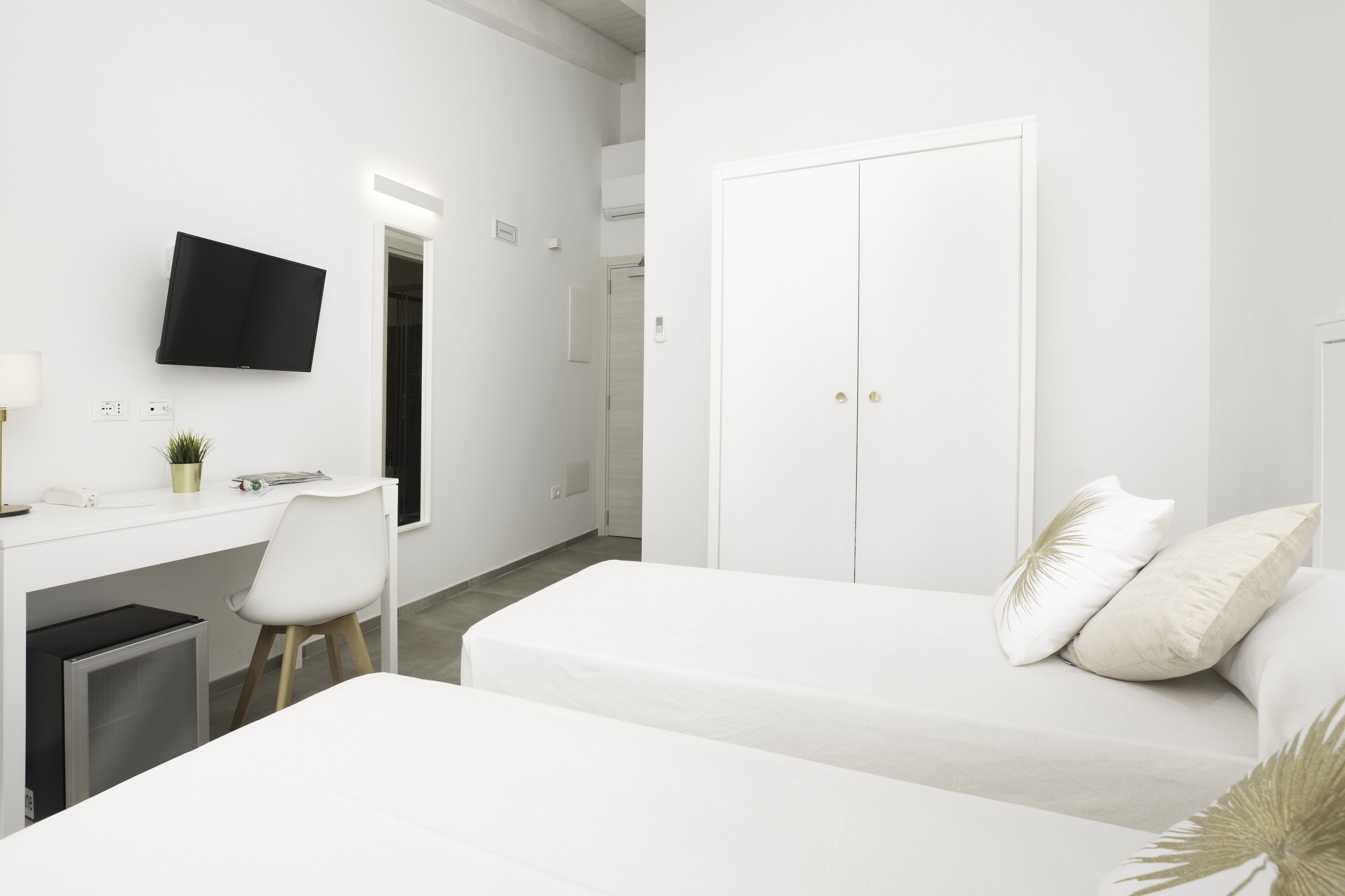 A modern hotel room with a white desk, a flat-screen TV, a bed with white bedding, and a wardrobe.