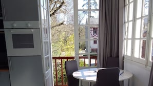 Une pièce compacte comprenant un coin cuisine, une table à manger avec des chaises, un canapé blanc moelleux et une vue sur un immeuble et des arbres, visible à travers une grande fenêtre.