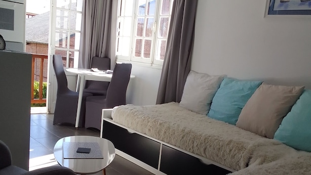 Un salon moderne avec un canapé, une petite table et un fauteuil. Il y a une fenêtre avec des rideaux, un tableau au mur et une vue sur l’extérieur.