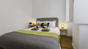 Una cama bien tendida con una manta gris y verde, dos mesitas de noche con lámparas y una lámpara colgante moderna.