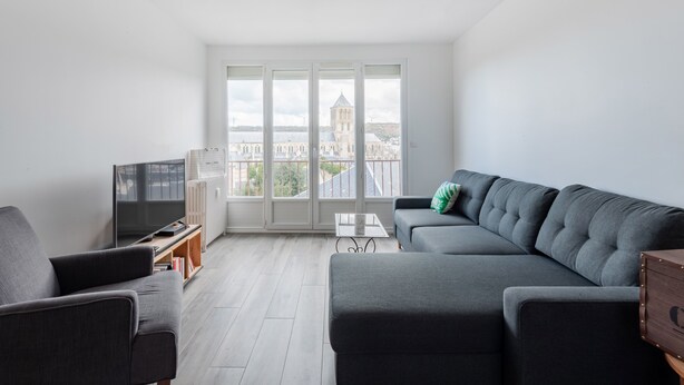 Una sala de estar moderna con un sofá gris, un televisor y vistas a un paisaje urbano a través de amplios ventanales.