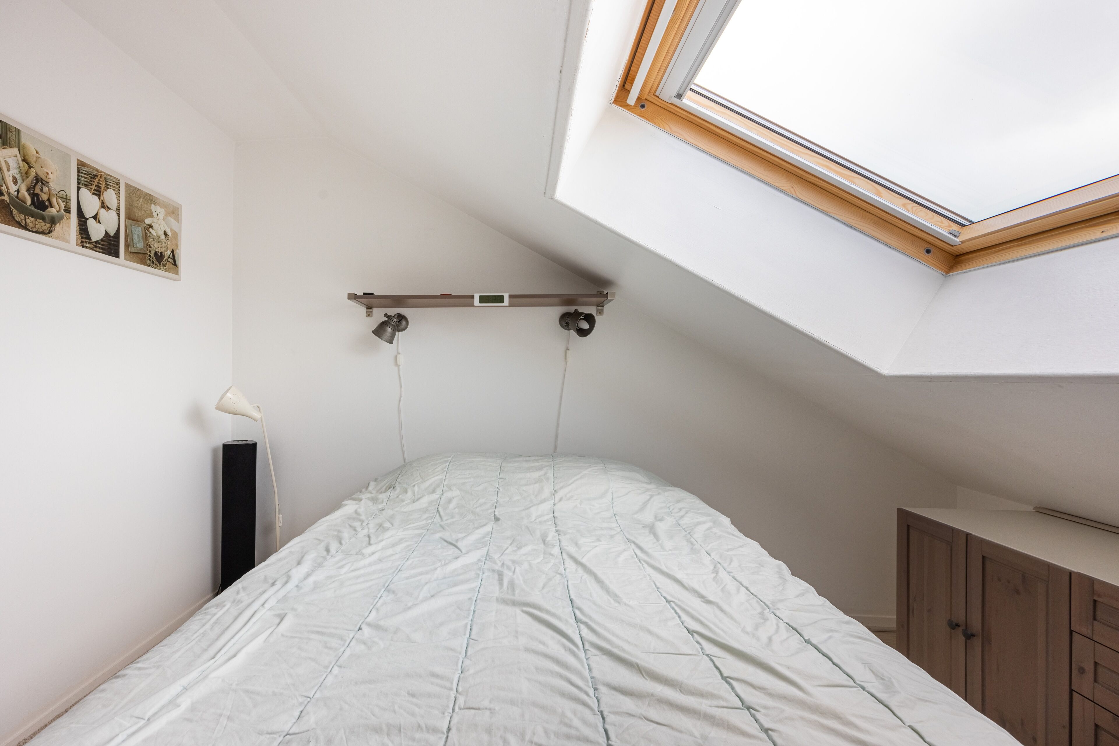 Een slaapkamer met dakraam, een bed, een bedlampje en een houten garderobe.