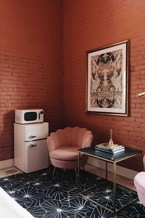 Ein Zimmer mit Backsteinwand, einem weißen Kühlschrank, einem rosafarbenen Stuhl, einem Glastisch mit Büchern und einem gerahmten Kunstwerk.