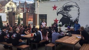 A group of people seated at wooden tables outdoors, with a mural of an eagle on a wall in the background.