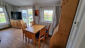 Ein Essbereich mit Holztisch und Stühlen, einem Fernseher auf einem Schrank und großen Fenstern mit Vorhängen.