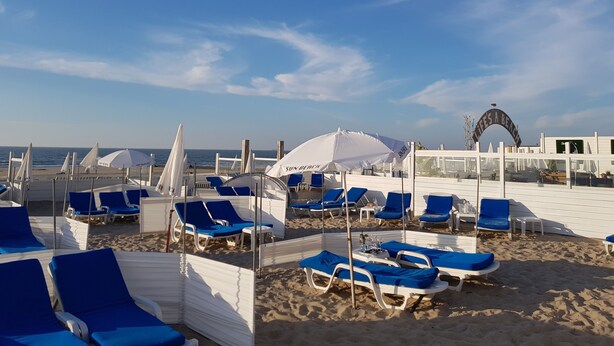 Ein Strand mit Reihen blauer Liegestühle und weißer Sonnenschirme, ein klarer Himmel und ein Schild mit der Aufschrift 'LIFE'S A BEACH'.