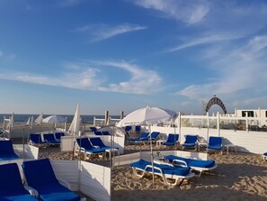 Une plage bordée de rangées de chaises longues bleues et de parasols blancs, un ciel dégagé et un panneau sur lequel on peut lire : « LA VIE EST UNE PLAGE ».