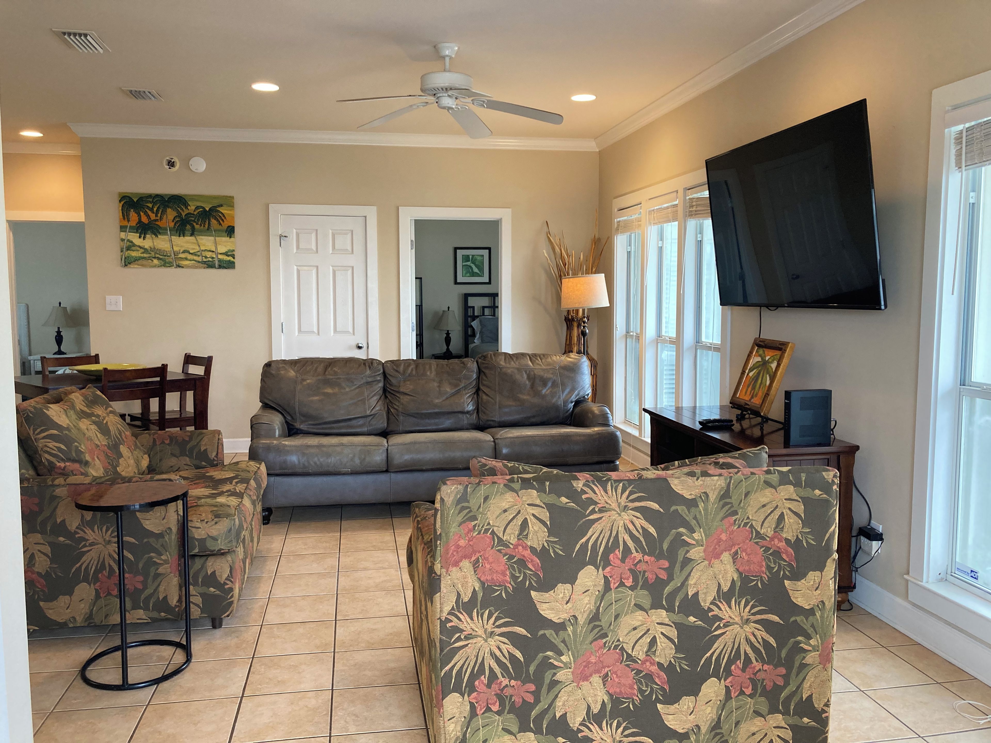 A living room with a sofa, two armchairs, a dining table with chairs, a ceiling fan, a TV mounted on the wall, and a window with blinds.