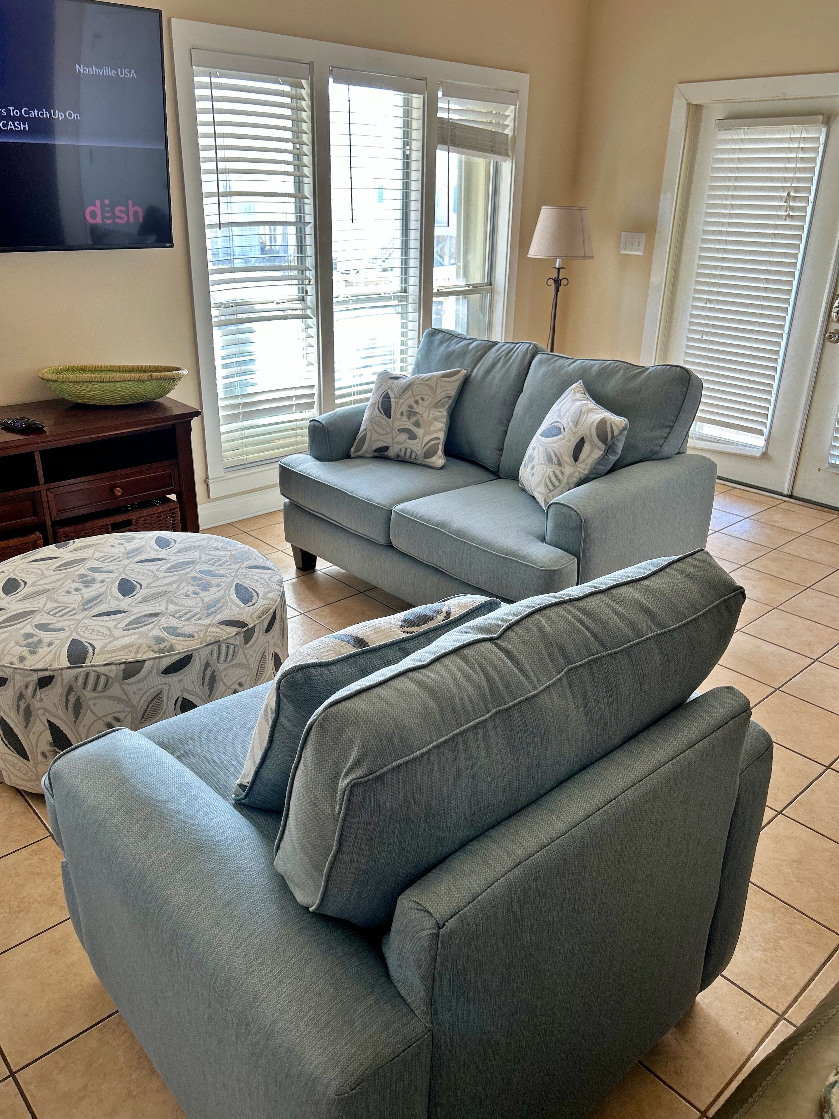 A living room with a grey sofa, patterned ottoman, and a television on the wall.
